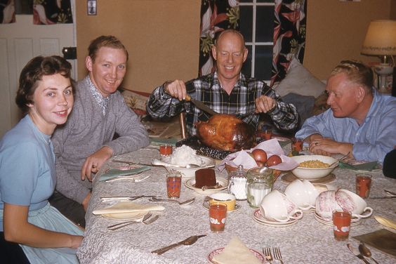 1950s vintage photo of a 1950s family sitting around the Thanksgiving dinner table getting ready to carve the turkey.
