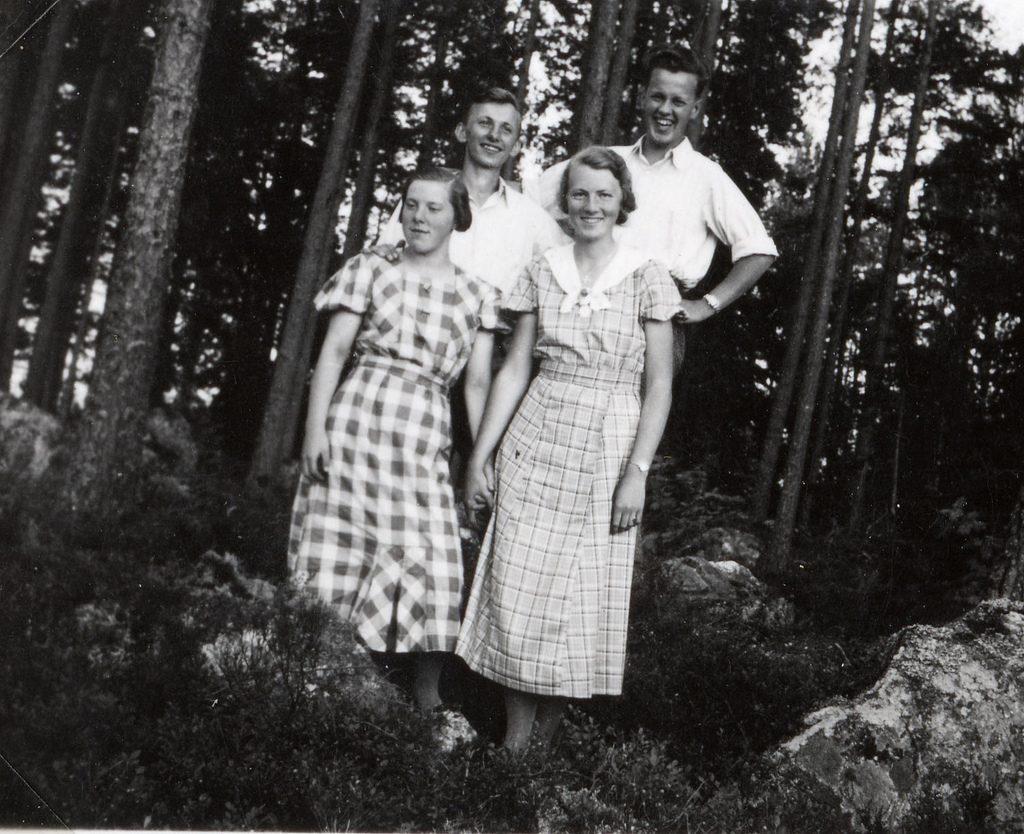 1930s vintage photo of two young women in 1930s dresses posing for a photo with two young men behind them in the woods. 