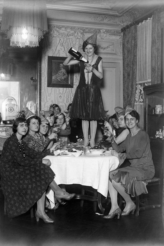 1920s New years eve party featuring a woman in 1920s dress standing on a table pouring champagne with other woman in 1920s dresses. 
