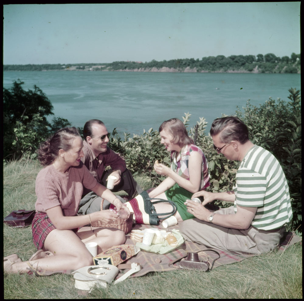 1950s vintage photo of  two couples having a picnic together beside Niagara Falls river in 1950s fashions. 