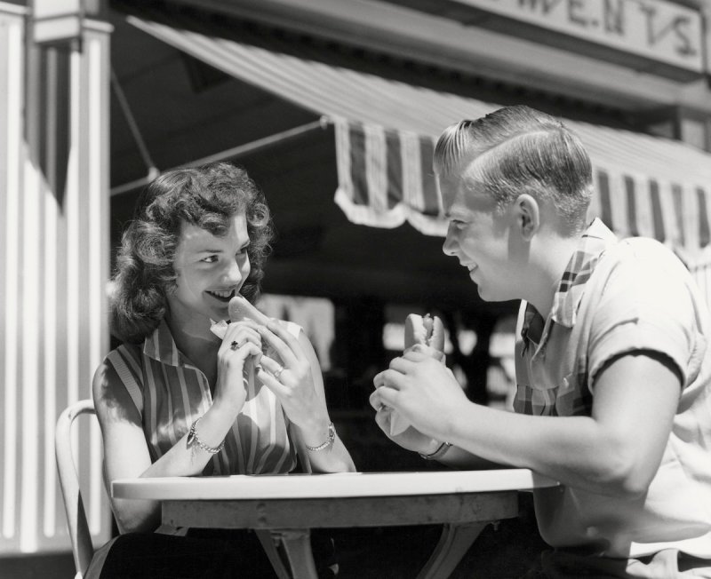 Teenage couple eating hotdogs outside at refreshment stand table, circa 1950 H. Armstrong Roberts—Retrofile/Getty Images