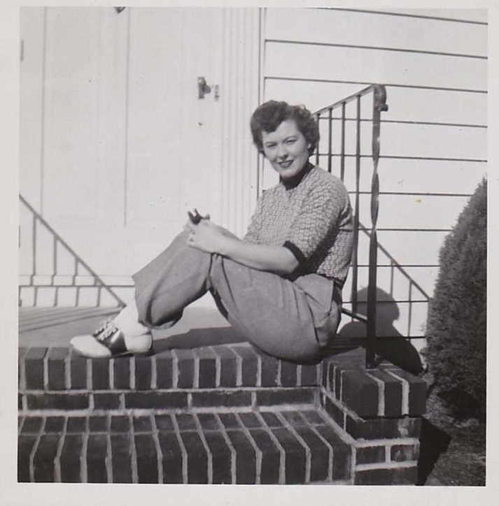 1950s Photo of a young woman in saddle shoes, pants and top with short 1950s vintage hairstyle sitting on her front step.