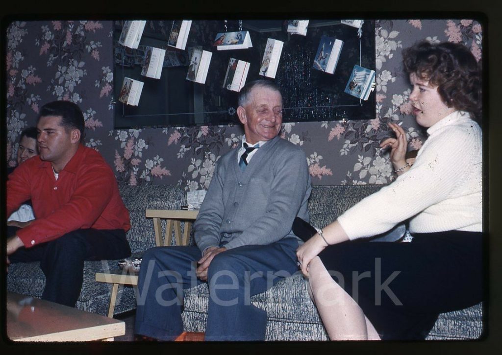 1960s vintage photo of people sitting in a living room in front of a floral wallpaper wall in 1960s clothing at Christmas time. 