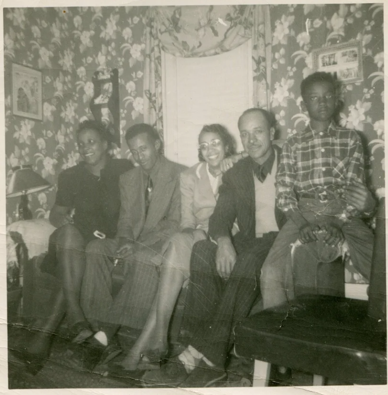 1950s vintage photo of a Black family posing together in their living room on their couch. The living room walls have floral wall paper and floral curtains. 
