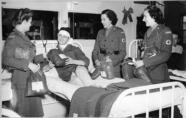 1940s Vintage Photo: Three CRCC (Canadian Red Cross Corps) Welfare Officers visit a convalescing soldier overseas (ca. 1943-45) while distributing “ditty bags” on a hospital ward.