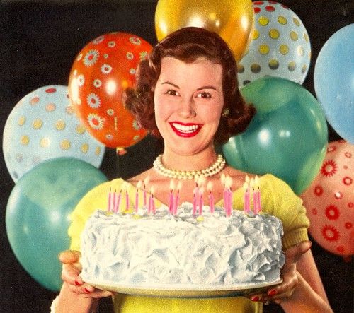 1950s vintage photo of a 1950s housewife holding a birthday cake with red lipstick, pearls and birthday balloons behind her