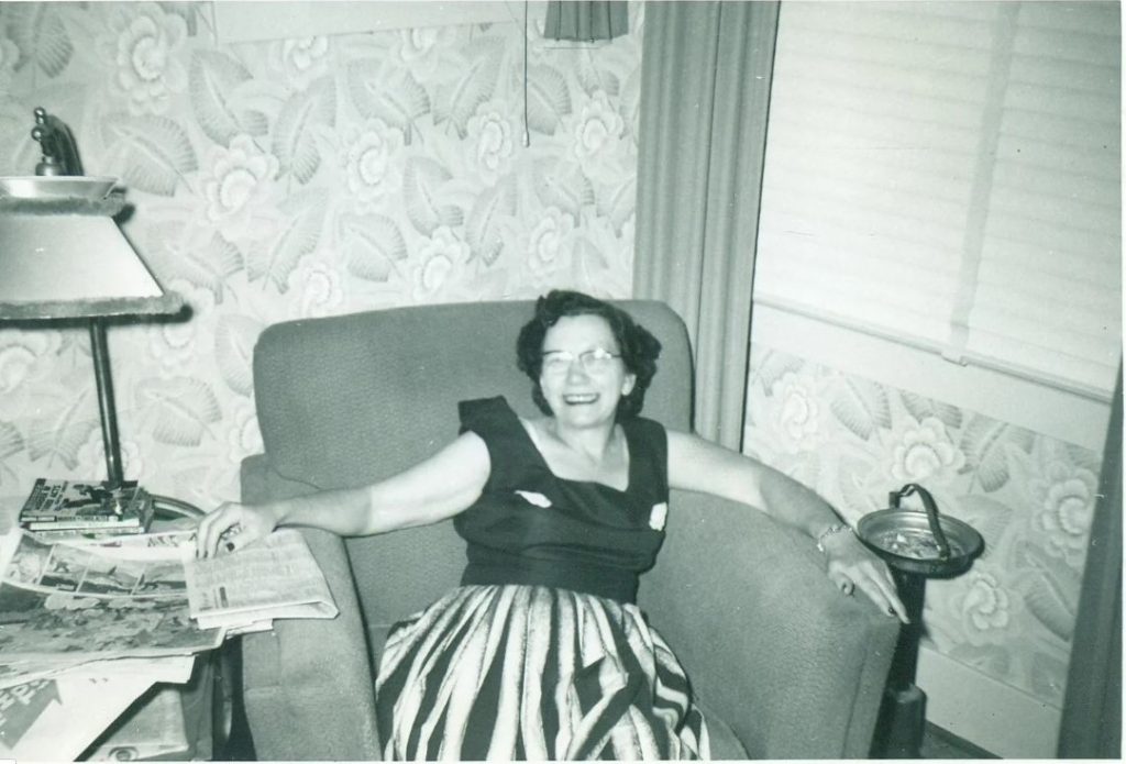 1950s vintage photo (November 1955) of an older woman in a 1950s dress sitting in chair in her living room with a 1950s hairstyle. Love the big smile on her face. 