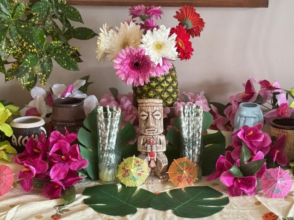tiki party decor tropical decor idea tropical tablescape by the Vintage Inn Blog. Pineapple vase is in the center and tiki mugs and faux flowers are used. 