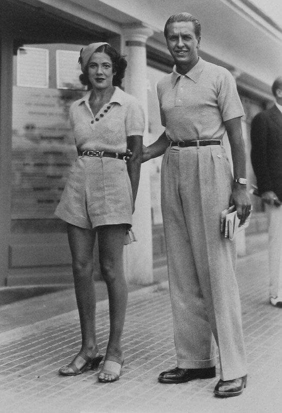 1930s vintage photo of a woman and man in 1930s fashions in Paris. The woman is wearing shorts, knit top and a hat and sandals. Our man is wearing pants and a golf shirt. 