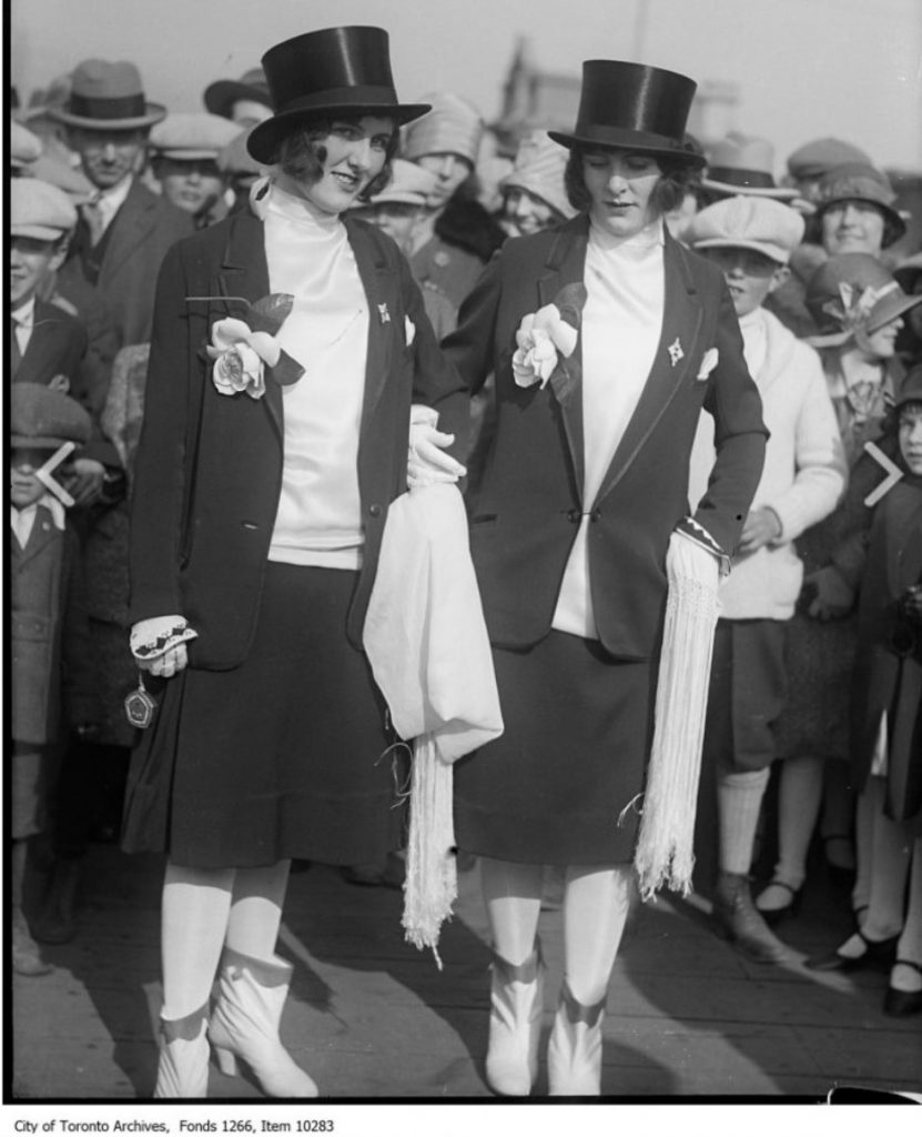 1920s vintage photo of two women at Toronto's Easter Parade wearing their own take on men’s tuxedos, complete with top hats as headwear. They’ve added a playful twist with matching white, cowboy-style boots and floral lapel pins.