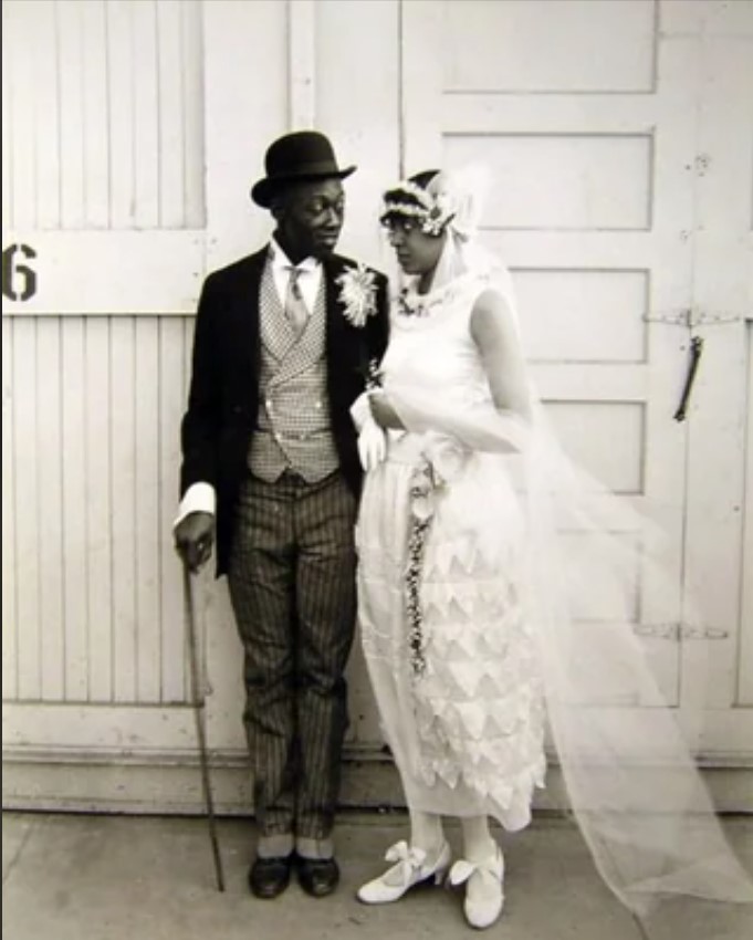 1920s vintage wedding photo of a Black bride and groom on their wedding day. Beautiful 1920s wedding dress and 1920s shoes. The groom is handsome in his bowler hat, floral brooch corsage and three piece suit. Very dapper. 