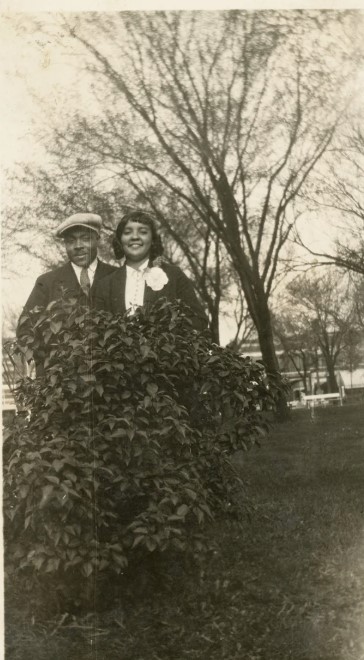 1930s or 1940s photo of a Black couple posing behind a bush. Our handsome gentlemen is sporting the popular 'Newsboy cap'.