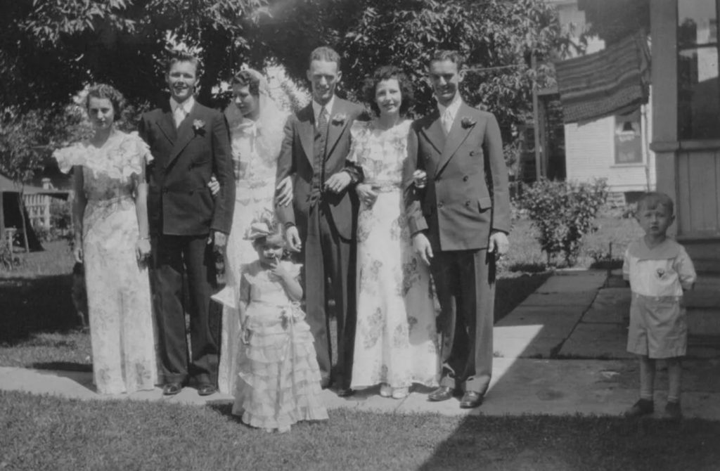 1930s vintage wedding photo from 1934 of bridesmaids in floral 1930s dresses, groomsmen in suits and the bride and groom posing with a flower girl and a little boy. 
