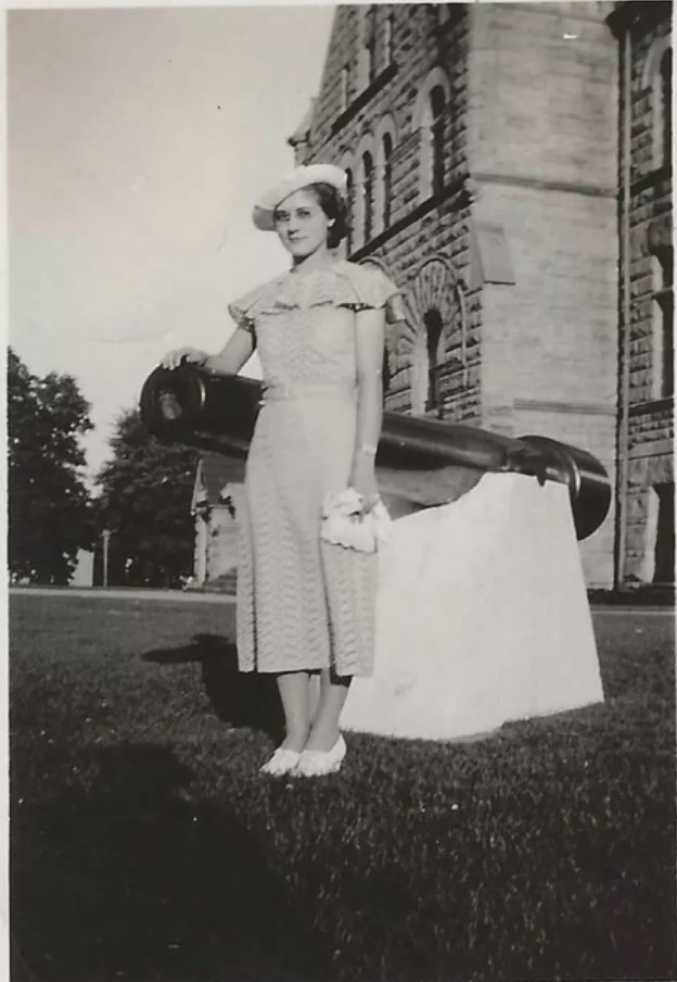 1930s vintage photo of a young woman in a 1930s dress, 1930s hat posing beside a cannon. She is also wearing glasses. Fantastic 1930s fashion.