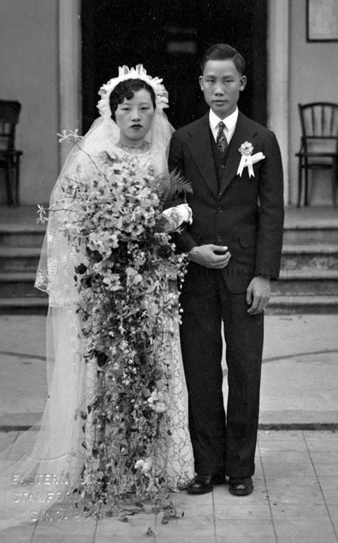 1920s or 1930s vintage photo of an Asian couple on their wedding day. The bride is stunning in a long veil and long wedding dress with a giant floral bouquet. The groom is in a simple suit. 