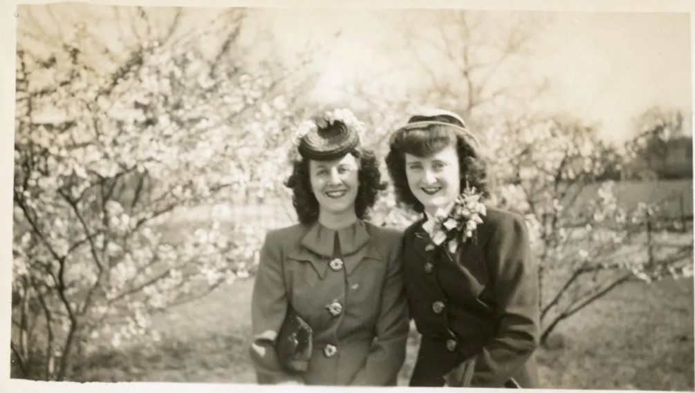 1940s vintage photo of two young women wearing 1940s hats posing in front of blooming trees during Easter time. The one woman is wearing a corsage and they both have 1940s hairstyles. 