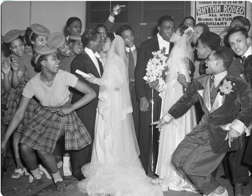 Black Love: 1940s vintage photo of a two Black couples after their wedding posing together for a kiss for the camera with the excited stylish groomsman and adorable bridesmaid (cute plaid skirts and matching hats)