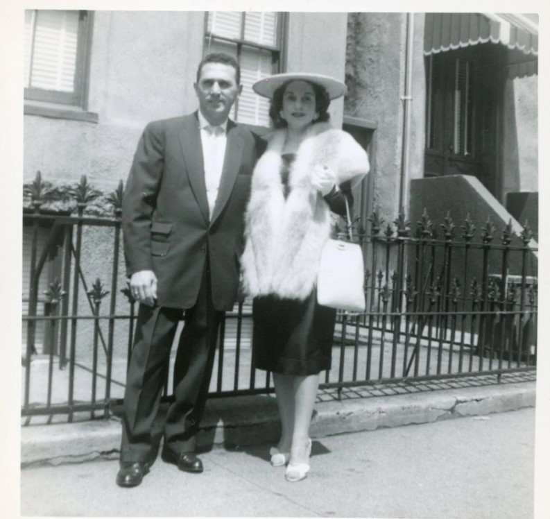 1950s photo / 1950s fashion of a stylish couple posing on the sidewalk. Our 1950s woman is sporting a fantastic platter hat, fur wrap over a black dress and a great purse. 