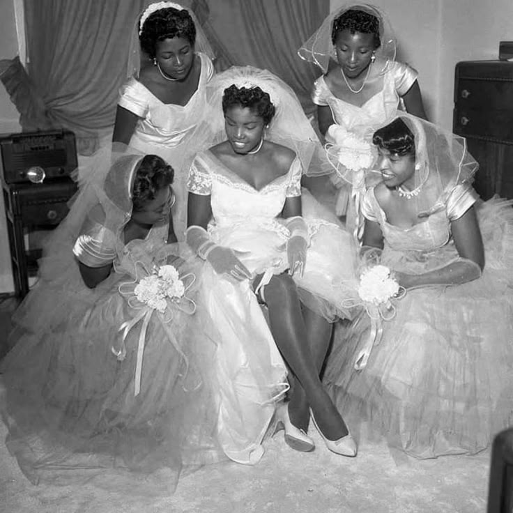 1950s vintage photo of a Black Bride surrounded by her bridesmaids as she puts on the garter under her 1950s wedding dress. 