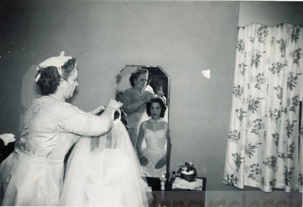 1950s vintage photo of a bride in a 1950s wedding dress having her mother put her veil on her head in this 1955 photo. 