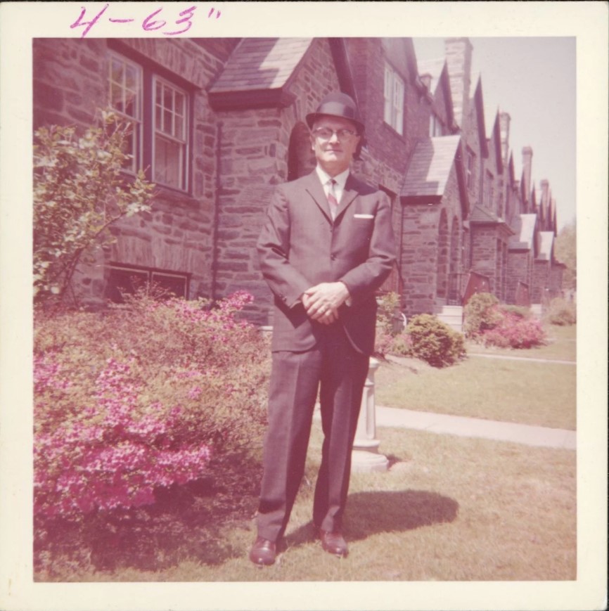 1960s vintage photo from 1963 of a Dapper Stylish Man in a Suit, Tie, bowler Hat posing by a flowering Shrub
