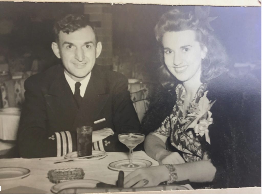 1940s vintage photo of a man in uniform dining with a pretty woman with a soft 1940s hairstyle with hair flower and corsage on her lapel. 1940s fashions. 